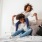 Lifestyle image of a mom holding her child on her shoulders while sitting on a bed 